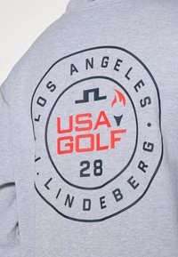 Grå hettegenser med rund grafisk design. Teksten sier "LOS ANGELES USA GOLF 28" i rødt og svart, akkompagnert av en flammegrafikk.