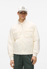 Giacca crema chiaro con cerniera, due tasche frontali e polsini a costine, abbinata a pantaloncini verde scuro e un cappello bianco con ricamo verde.