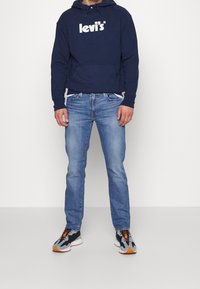 Námořnická modrá mikina s bílým logem "Levi's", v kombinaci s světle modrými džínami a černými teniskami s oranžovými detaily. Model stojí čelem.