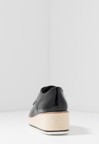 Chaussure en cuir verni noir avec un talon compensé, présentant un design en jute tissé. Surface lisse avec bords arrondis et légère brillance.