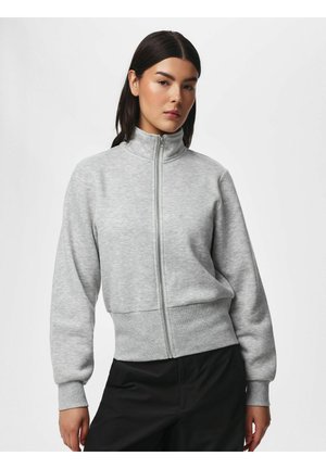 Vrouw met donker haar die een lichtgrijze zip-up trui met hoge kraag en zwarte broek draagt, staand tegen een effen witte achtergrond.