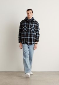 Rutet skjorte i svart, blått og hvitt med to brystlommer, sammen med lyseblå jeans og hvite sneakers, mot en nøytral bakgrunn.