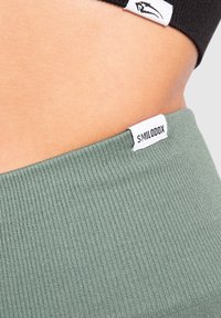 Ingrandimento della vita di una persona che indossa una fascia nera e un tessuto a coste verde con un'etichetta bianca "SMILODOX".