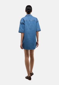 Robe chemise en denim oversized bleu avec des manches courtes, coupe droite et col classique, présentant des coutures contrastantes et un ourlet arrondi.