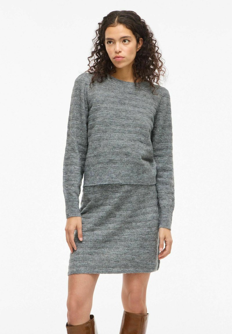 Conjunto de suéter y falda de punto gris con un patrón texturizado. El suéter tiene mangas largas y un cuello redondo. Botas altas marrones.