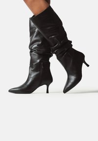 Cizme din piele negre, până la jumătatea gambei, cu fermoar lateral, vârf ascuțit, cu un șir lung și toc kitten, purtate cu picioarele încrucișate pe un fundal alb.