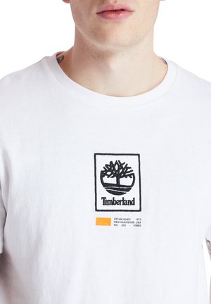 Muž v bílem tričku Timberland s černým logem stromu a nápisem na hrudi, viditelné tetování na krku, oříznutý u brady a ramen.