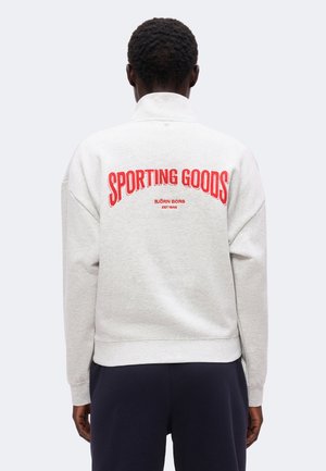 Personne portant un sweat-shirt gris clair avec un texte rouge ""SPORTING GOODS BJÖRN BORG EST 1956"" dans le dos et un pantalon foncé, vue de dos.