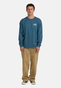 Mężczyzna stoi twarzą do przodu, ubrany w niebieską koszulę z długim rękawem z małym logo, beżowe spodnie i czarne sneakersy na białym tle.