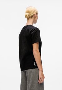 Blonde woman with hair in a bun wears a plain black t-shirt and gray pants, standing with her back turned on a white background.