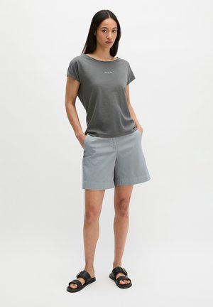 Frau mit langen dunklen Haaren steht mit den Händen in den Taschen, trägt ein graues T-Shirt mit kurzen Ärmeln, hellblaue knielange Shorts und schwarze Slip-on-Sandalen.