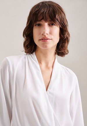 Blusa bianca in stile avvolgente realizzata in tessuto leggero con maniche lunghe e un dettaglio a bottone singolo presso il collo. Texture morbida, aspetto liscio.