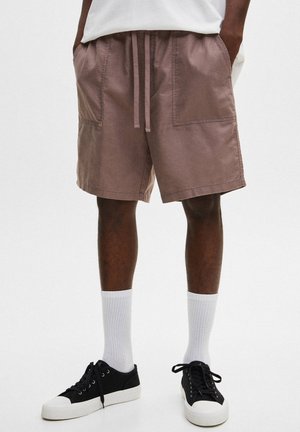 Homme portant un short décontracté mauve avec taille à cordon, chaussettes blanches montantes, baskets noires et blanches, et t-shirt blanc, debout les mains dans les poches.