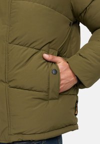 Hånd i lommen på en olivengrøn polstret jakke med sort trykknap og lille sort og orange etiket på jakkens kant.