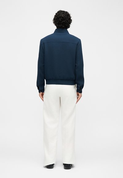 Osoba s kučeravými vlasmi, viditeľná zozadu, v tmavomodrej bunde, bielych nohaviciach s širokými nohavicami a čiernych topánkach na jednoliatom pozadí.