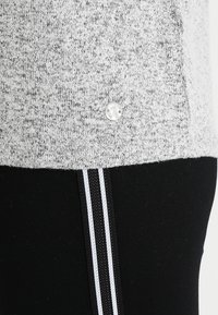 Un pull en tricot gris chiné avec un ourlet et une petite épingle en argent rond, porté sur un pantalon noir avec des rayures blanches sur les côtés.