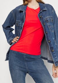 Jeansjacka med två bröstfickor, blå i färgen, över en åtsittande röd t-shirt. Modellen bär blå jeans, vilket visar en avslappnad outfit.