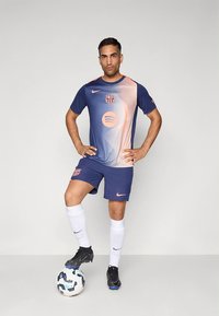 Nike Barcelona fotbollskit i marinblått och gradient orange. Innehåller logotyp, Spotify-ikon, shorts och svarta fotbollsskor. Står på en fotboll.