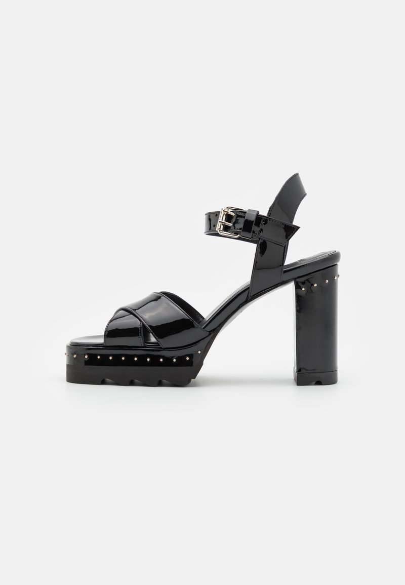 Sandalias de plataforma de charol negro con correa ajustable en el tobillo, puntera redondeada y acentos de tachuelas a lo largo de la base. Tacón en bloque grueso.
