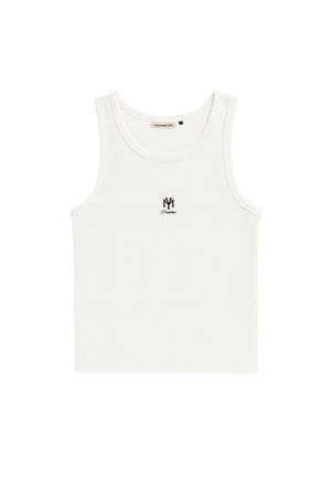 Joven con camiseta blanca sin mangas y gorra bicolor, sosteniendo una camisa azul a rayas sobre su hombro sobre un fondo liso.