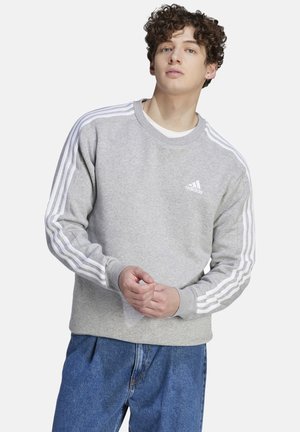 Felpa grigia con logo Adidas bianco e tre strisce bianche sulle maniche. Presenta un collo rotondo e polsini a costine, abbinata a jeans blu.