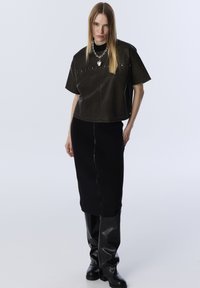 Donna che indossa un top in pelle nera a maniche corte, una gonna midi a costine nere con zip frontale, stivali al ginocchio in pelle nera e una collana a forma di cuore in argento.