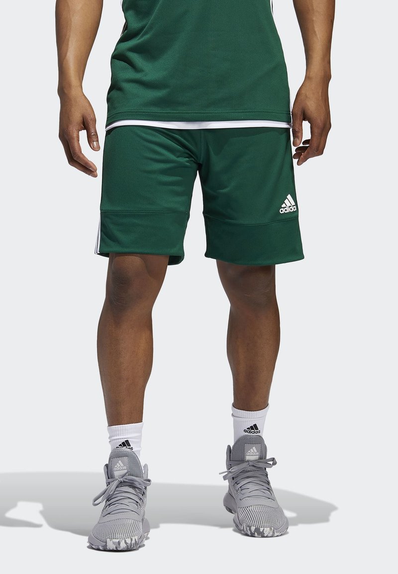 Grüne Sportshorts aus atmungsaktivem Material mit weißen Akzenten und einem Dreistreifen-Design. Kombiniert mit grauen Schuhen und weißen Socken.