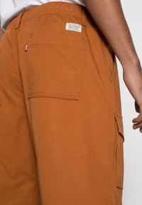 Braune Cargo-Hosen mit mehreren Taschen, verstellbarem Bund und einem Etikett. Der Stoff wirkt robust und hat eine glatte Textur.