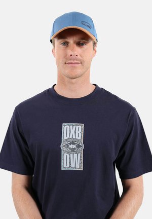 Homme portant un t-shirt bleu marine avec un logo vertical « OXBOW » et une casquette de baseball Oxbow bleu et beige, debout devant un fond blanc.