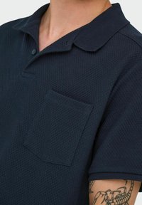 Navy blue polo shirt with a textured fabric, button placket, and a breast pocket. Short sleeves with a classic collar design.