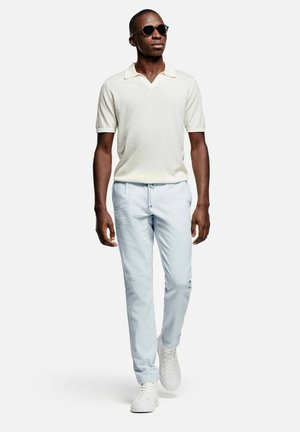 Homme portant des lunettes de soleil, un polo blanc à manches courtes, un pantalon bleu clair avec cordon de serrage et des baskets blanches, marchant en avant sur un fond uni.