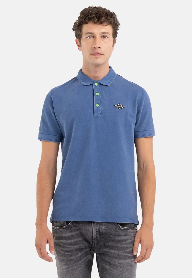 Camisa polo azul de algodón con mangas cortas, botones verdes y un logo de diamante negro en el pecho. El diseño es simple y relajado.