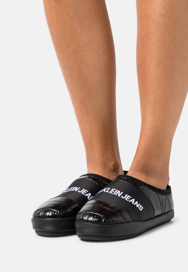 Personne portant des chaussures noires matelassées à enfiler avec l'inscription "KLEIN JEANS" en blanc sur le dessus, debout sur un fond neutre.