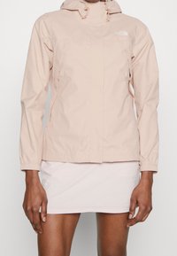 Giacca impermeabile rosa chiaro con zip frontale, cappuccio regolabile, polsini elasticizzati e due tasche laterali. Tessuto liscio, branding minimale sul petto.