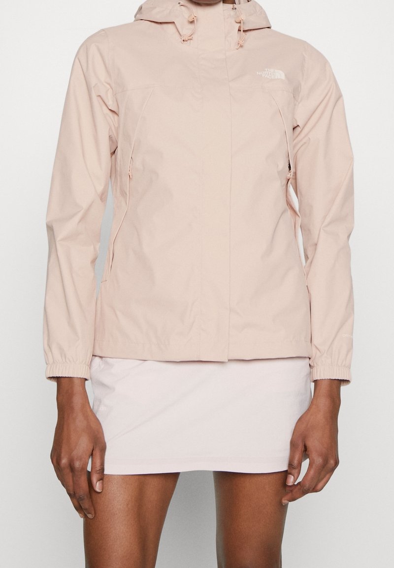 Giacca impermeabile rosa chiaro con zip frontale, cappuccio regolabile, polsini elasticizzati e due tasche laterali. Tessuto liscio, branding minimale sul petto.