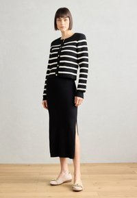 Svartvitt randig kofta med guldknappar, kombinerad med en svart ribbad klänning i ankellängd med en sidoklyvning och beigea platta skor.