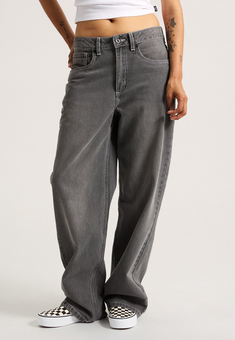 Personne portant un jean gris délavé ample, un top court blanc et des chaussures slip-on à carreaux noir et blanc, debout devant un fond uni.