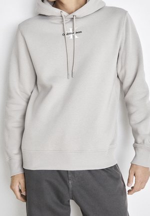 Mand iført en lys beige Calvin Klein hoodie med snore og mørke grå bukser, der står foran en ensfarvet baggrund.