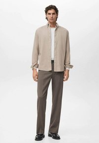 Camisa de botones color beige sobre una camiseta blanca, combinada con pantalones marrones y zapatos negros. La camisa tiene las mangas arremangadas y un corte relajado.