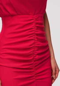 Gros plan de la taille d'une robe rouge froncée avec des coutures latérales visibles et un bras partiellement visible sur un fond neutre.