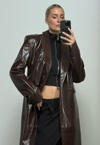 Γυναίκα που βγάζει selfie στον καθρέφτη φορώντας ένα γυαλιστερό σκούρο καφέ δερμάτινο παλτό, μαύρο cropped τοπ με φερμουάρ και μαύρο παντελόνι με φόντο μονόχρωμο.