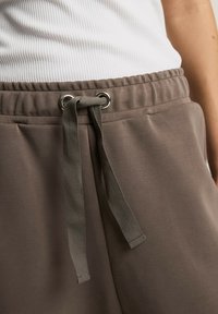 Brązowe spodnie z elastycznym pasem, wyposażone w sznurek z metalowymi oczkami, gładką fakturą oraz bocznymi kieszeniami. Zbliżenie na szczegóły.