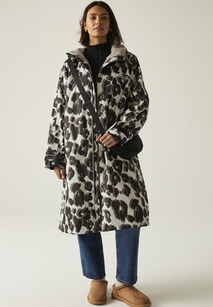 Kobieta ubrana w czarno-biały płaszcz w print lamparta o długości do połowy uda, niebieskie dżinsy, beżowe pantofle wsuwane i czarną torebkę na ramię, stojąca na jednolitym tle.