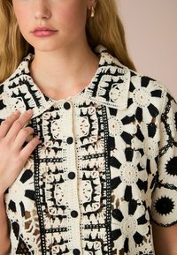 Hæklede bluse i creme og sort med blomster- og geometriske mønstre, korte ærmer og knaplukning fortil. Textile detaljer kan ses.