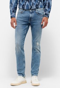 Svetlo modré džínsové nohavice so slim fit strihom, so zničením na kolenách a piatimi vreckami; spárované s bielymi teniskami.