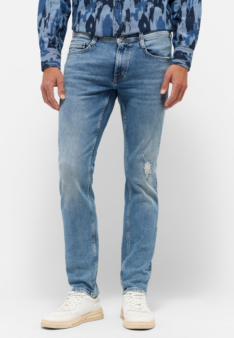 Svetlo modré džínsové nohavice so slim fit strihom, so zničením na kolenách a piatimi vreckami; spárované s bielymi teniskami.