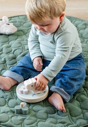 Jouet d'empilage d'animaux en bois avec des couleurs pastel; comprend une base circulaire et des pièces amovibles. L'enfant est assis sur un tapis matelassé vert.