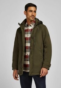 Olijfgroen gewatteerd jack met capuchon, voorzien van ritszakken en een gladde buitenkant. Gekozen boven een meerkleurig geruit flanellen overhemd.