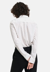White long-sleeve shirt with black polka dots, featuring a collar and buttoned cuffs, paired with black trousers. Back view.