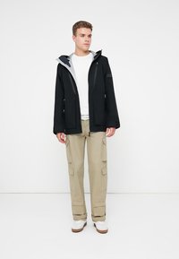 Must värviga veekindel jakk, millel on lukuga esikülg, heledat halli voodrit ja kaks lukuga taskut, millele on lisatud beežid cargo-püksid ja valged spordijalanõud.